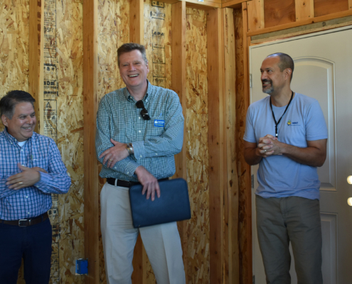 Tom Collishaw, wearing a green checkered shirt, stands inside an unfinished home alongside HAC CEO David Lipsetz and others. They are smiling and laughing as they discuss housing development. The background features exposed wooden framing and a closed white door.